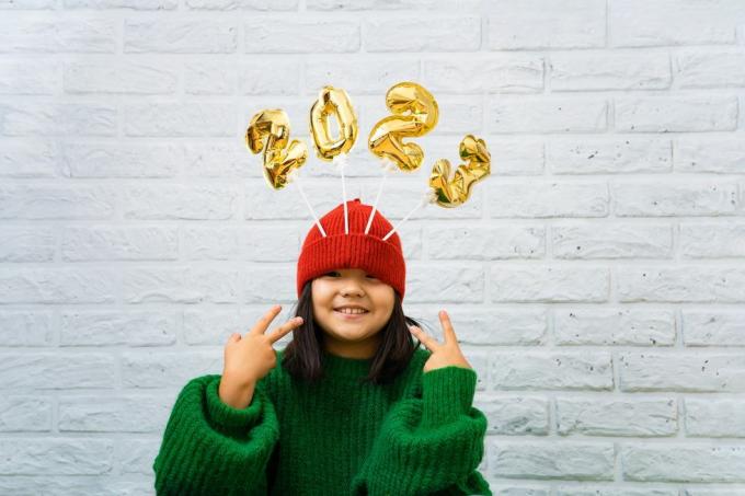 Fröhliches asiatisches Mädchen in einem grünen Pullover, Nahaufnahme mit goldenen Luftballons mit den Zahlen 2023 in einem Hutkopierraum, weißer Backsteinwandhintergrund, Mädchen blickt in die Kamera und lächelt