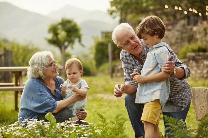 Großeltern unterhalten sich mit zwei kleinen Kindern im Garten, im Hintergrund Berge und Haus