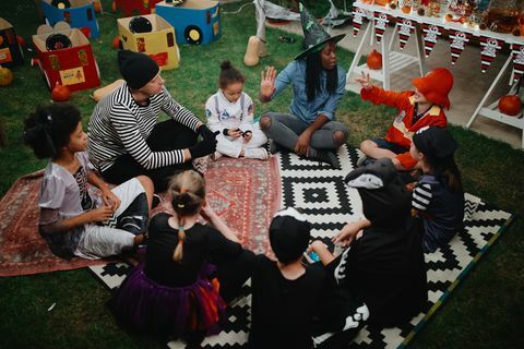 Eltern erzählen Kindern auf einer Halloween-Party im Freien Gruselgeschichten. Sie sitzen mit gekreuzten Beinen auf Teppichen im Gras. Ein Elternteil trägt einen Hexenhut