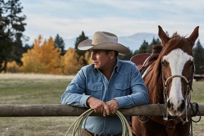 kevin costner in yellowstone neben einem pferd, das sich mit einem seil in den händen an einen zaun lehnt und ein verblichenes blaues jeanshemd und einen beigen cowboyhut trägt