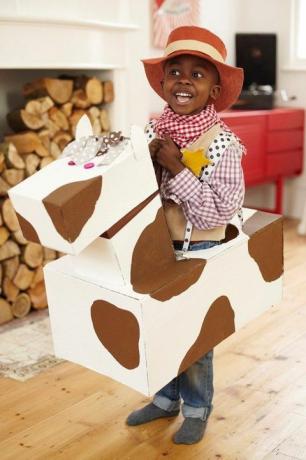 kleiner Junge als Cowboy verkleidet mit Cowboyhut und kariertem Hemd und Bandana mit einem Papppferd um die Taille
