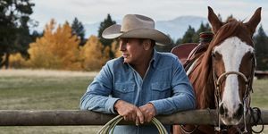 kevin costner in yellowstone neben einem pferd, das sich mit einem seil in den händen an einen zaun lehnt und ein verblichenes blaues jeanshemd und einen beigen cowboyhut trägt