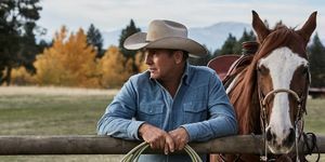 kevin costner in yellowstone neben einem pferd, das sich mit einem seil in den händen an einen zaun lehnt und ein verblichenes blaues jeanshemd und einen beigen cowboyhut trägt