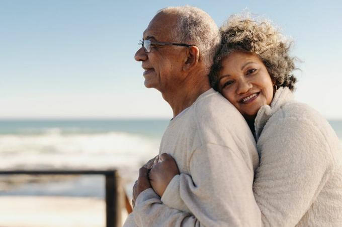 Romantische ältere Frau lächelt in die Kamera, während sie ihren Mann am Meer umarmt. Zärtliches älteres Paar genießt es, nach der Pensionierung eine schöne gemeinsame Zeit zu verbringen