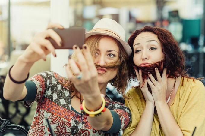 Zwei Freundinnen halten sich Haare vors Gesicht, um auszusehen, als hätten sie Bart und Schnurrbart, und machen ein Selfie davon