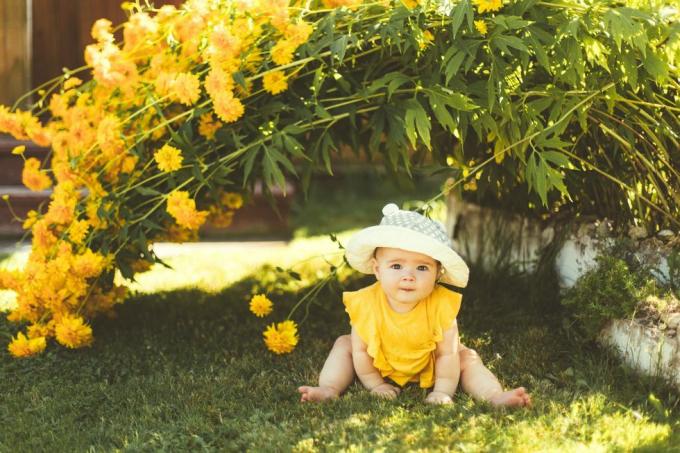 Ein kleines Mädchen mit Sonnenhut sitzt im Garten unter einem großen gelben Blumenstrauch