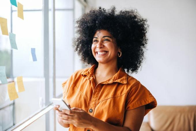 Glückliche schwarze Geschäftsfrau lächelt, während sie in einem Kreativbüro ein Smartphone benutzt. Fröhliche Unternehmerin sendet eine SMS, während sie neben einer Glaswand mit Haftnotizen steht