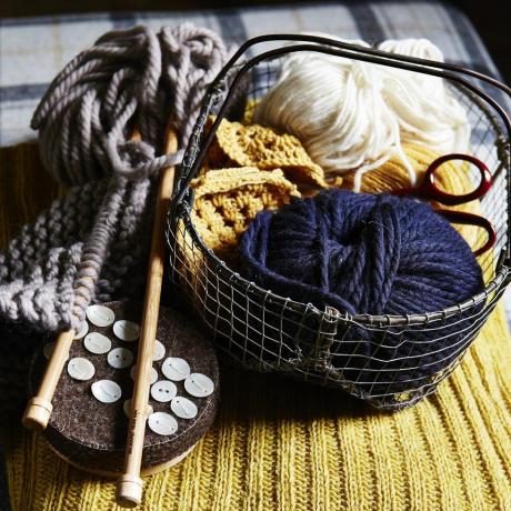 Knäuel aus blauer und weißer Wolle in einem kleinen Drahtkorb, Stricknadeln aus Holz, Knöpfe, auf gelbem geripptem Strickstoff platziert, Textur