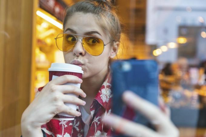 Junge Frau draußen mit Sonnenbrille macht ein Selfie und trinkt einen Kaffee zum Mitnehmen