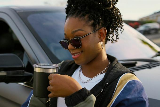 Izzys Charakter steht neben einem Lincoln-Geländewagen und trinkt eine Tasse Kaffee. Er trägt eine Sonnenbrille
