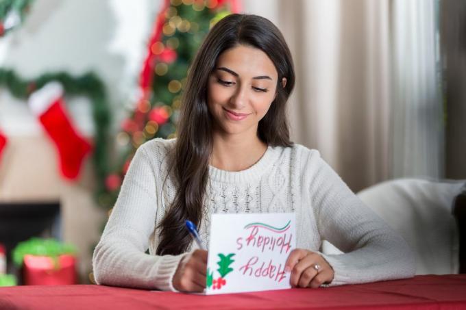 Eine lächelnde junge Frau sitzt an einem Tisch in ihrem dekorierten Wohnzimmer und blickt konzentriert nach unten, während sie einen Brief an eine Freundin zu Weihnachten schreibt