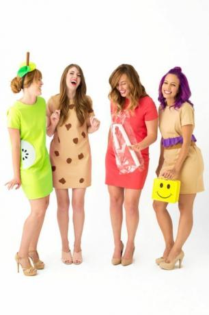 vier lachende Frauen in kurzen Kleidern, die als "Lunch Ladies" verkleidet sind, eine mit einer gelben Smiley-Lunchbox