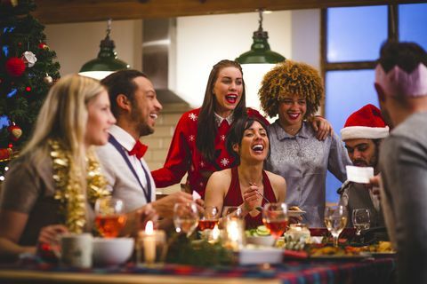 Eine Gruppe von Freunden, die lachen, während sie um einen Tisch sitzen und zu Weihnachten essen und trinken, ein Mann liest einen Witz von einer Weihnachtscrackerparty, Hüte und Weihnachtspullover werden getragen